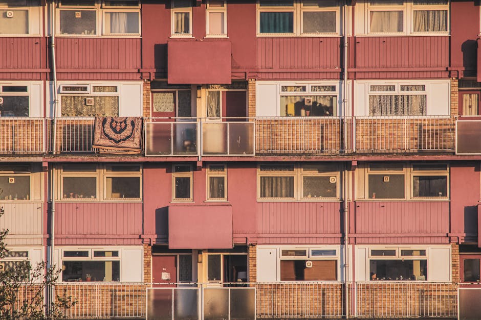 A multi-storey residential building with a pink exterior, featuring several rows of windows and balconies. The balconies are enclosed with metal and glass railings, some with visible items such as a p